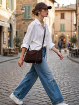 A woman walking on a cobblestone street wearing a white button-down shirt, wide-leg blue jeans, white sneakers, and a dark brown baseball cap, with a dark brown crossbody bag slung across her body.