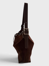 A dark brown faux suede shoulder bag with a wide adjustable shoulder strap and a silver-tone buckle, shown from a side profile view against a light gray background.