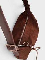 A close-up of a brown faux suede crossbody bag with a matching leather strap, silver zipper, and metal hardware against a light gray background.