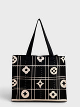 A black tote bag with cream-colored geometric and floral patterns, featuring two black shoulder straps, set against a light gray background.