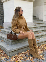 A woman sits on stone steps outdoors wearing a tan suede-like belted jacket, white shorts, and matching tan over-the-knee boots, with a brown handbag beside her.