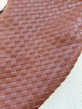 Close-up of a cognac shade brown woven leather bag surface showing the texture and pattern of the interlaced straps on a light beige background.