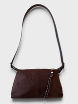 A dark brown faux suede shoulder bag with a matching braided leather tassel and a thin dark strap, shown against a light gray background and shown with a long crossbody strap.